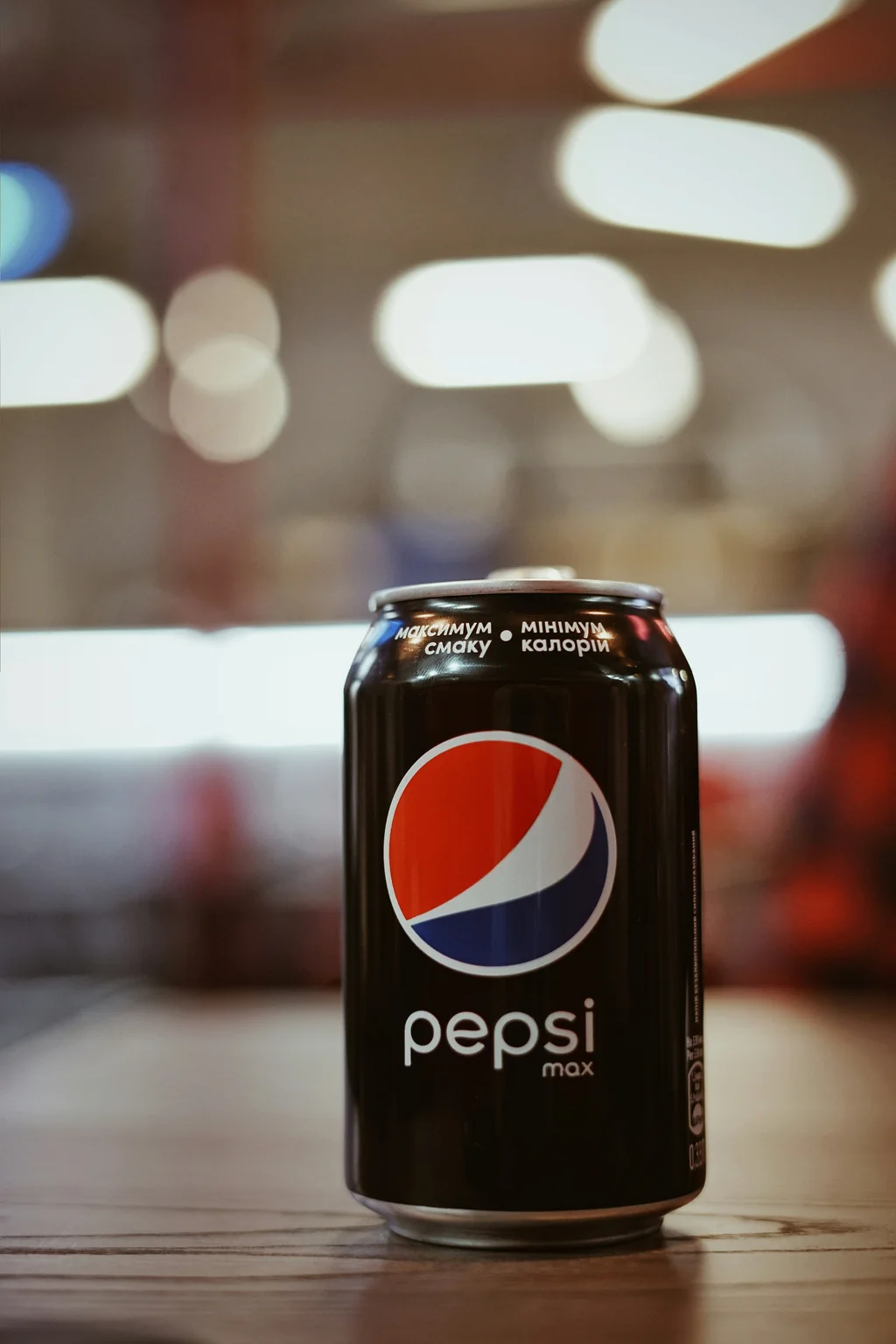 A close-up of a Pepsi Max can positioned centrally on a textured wooden table. The can features a glossy finish with the iconic red, white, and blue logo prominently displayed. The background offers a bokeh effect, with soft, circular light spots hinting at a casual setting, perhaps a café or gathering space. The warm lighting enhances the colors, creating a vibrant yet relaxed mood. Focused on the can, the shot utilizes a shallow depth of field to emphasize the subject while softly blurring the environment.